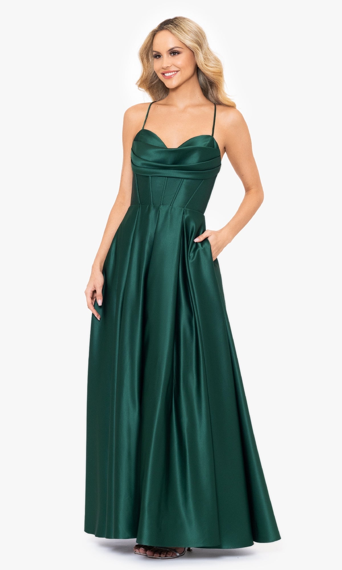 A woman models the Blondie Nites 3946BN Long A-Line Prom Dress, an emerald green satin gown with a corset-style waist and thin straps, standing against a white background.