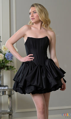 A woman in a black Colors Dress 3953 short homecoming dress, featuring a strapless corset bodice and ruffled skirt, stands indoors near a table with a floral arrangement.