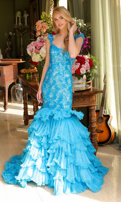 A woman stands indoors wearing the Long Prom Dress 39551 by Ava Presley, a bright blue off-the-shoulder mermaid gown with a layered ruffle skirt; flower arrangements and a guitar are visible in the background.