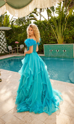 A woman in the Ava Presley 39553 Feather Long Prom Ball Gown, featuring a bright blue off-the-shoulder design and beaded bodice, stands poolside with lush greenery and patio furniture in the background.
