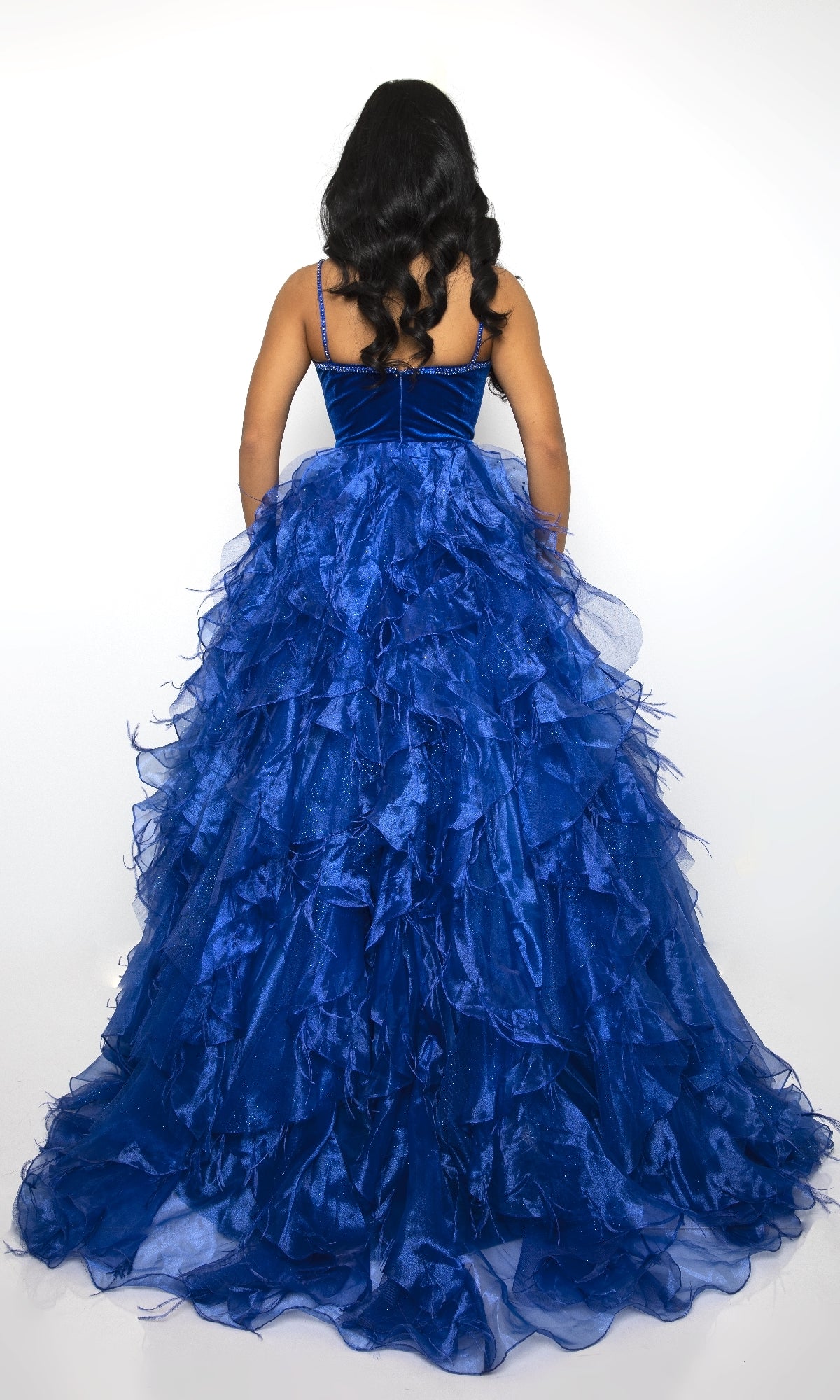 A person with long, wavy hair is shown from the back wearing the Long Ruffled Prom Ball Gown: Ava Presley 39554, a sleeveless blue floor-length dress with a voluminous ruffled skirt, against a white background.