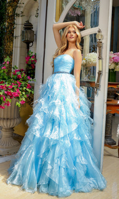 A young woman wearing the Ava Presley 39555 Strapless Ruffle Prom Gown stands by a glass door, surrounded by flowers and outdoor decor, her elegant A-line gown flowing around her.