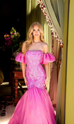 A woman stands indoors near a window with green curtains, wearing the Ava Presley 39570 puff-sleeve long prom dress. A guitar and flowers are visible in the background.