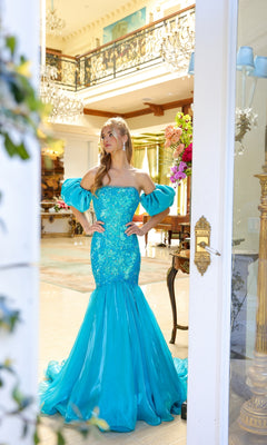 A woman in the Ava Presley 39570 Puff-Sleeve Long Prom Dress poses indoors near an open door, hands on hips, in an elegantly decorated room.