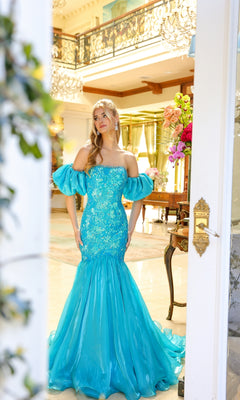 A woman stands indoors near a doorway, surrounded by ornate decor and flowers, wearing the Ava Presley 39570 puff-sleeve long prom dress in bright blue with an embellished bodice.