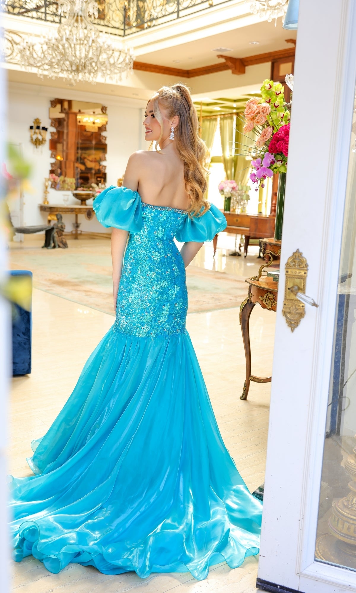 A woman wearing the Ava Presley 39570 Puff-Sleeve Long Prom Dress with an embellished bodice stands indoors, facing away, in a well-lit, elegant room decorated with flowers and chandeliers.