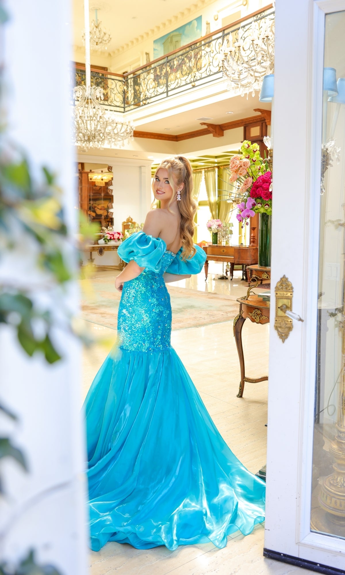 A woman wearing the Ava Presley 39570 puff-sleeve long prom dress smiles in an elegant, well-lit room adorned with chandeliers and floral arrangements.