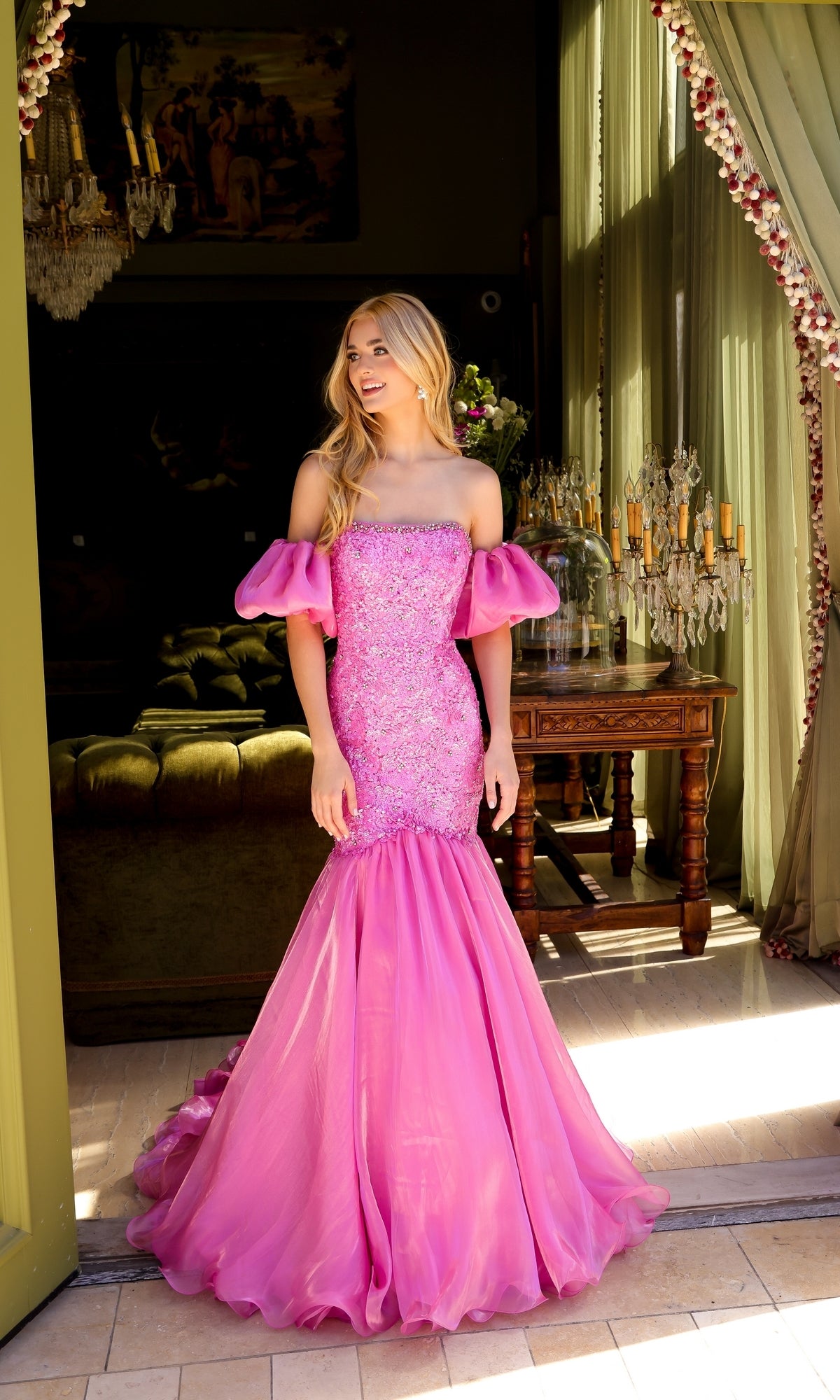 A woman stands in a doorway wearing the Ava Presley 39570, a bright pink puff-sleeve long prom dress with an off-shoulder mermaid silhouette and an embellished floral bodice.