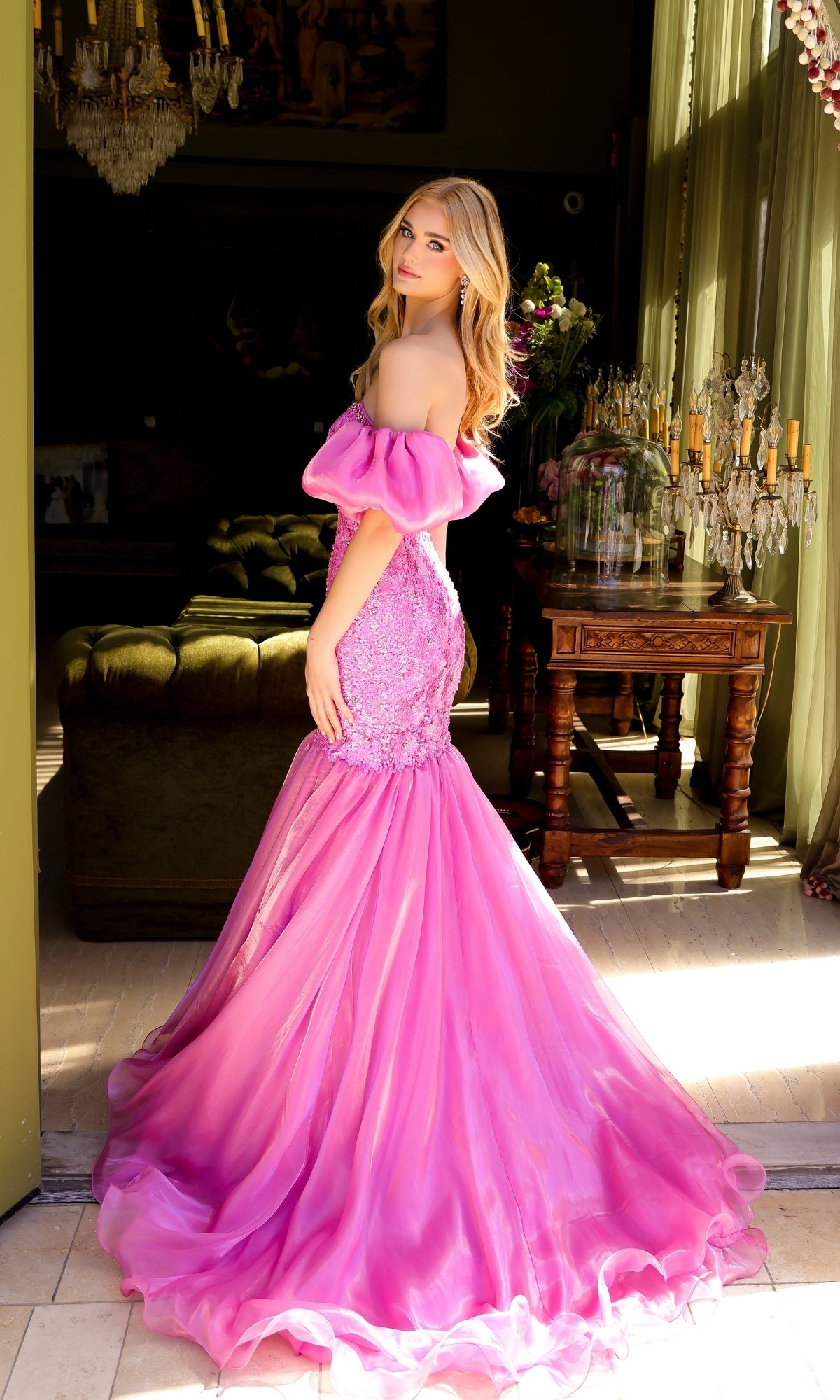 A woman in the Puff-Sleeve Long Prom Dress: Ava Presley 39570 stands indoors by a sunlit window, looking over her shoulder in a pink, off-the-shoulder mermaid dress with an embellished bodice.