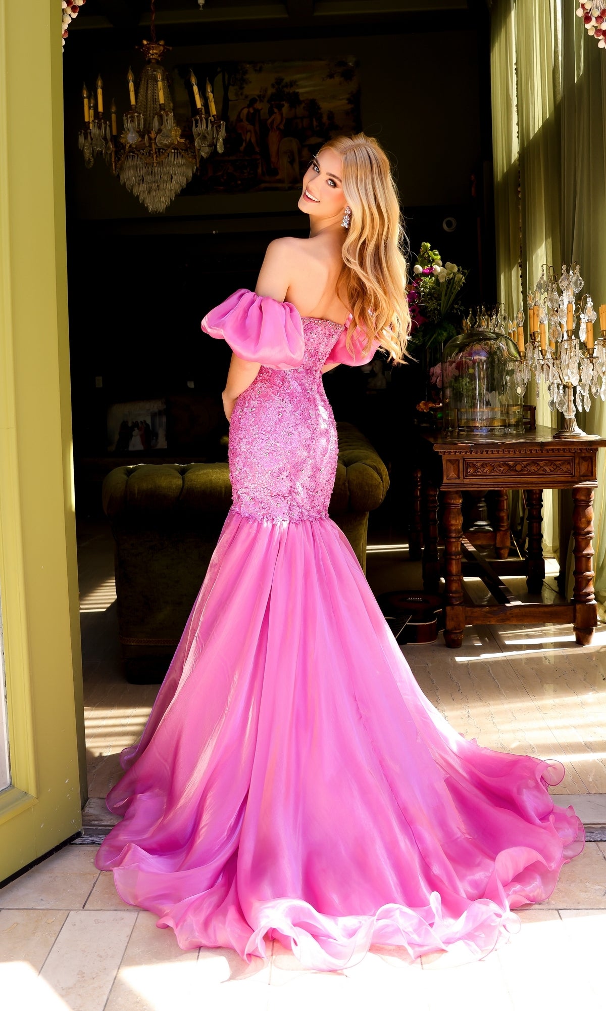 Wearing the Ava Presley 39570 puff-sleeve long prom dress with an embellished bodice, a woman stands indoors, facing away and glancing over her shoulder as sunlight pours through tall windows.