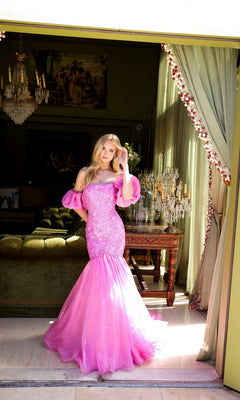 A woman stands in a doorway wearing the Ava Presley 39570 Puff-Sleeve Long Prom Dress; the fitted pink gown features off-the-shoulder ruffled sleeves and a tulle mermaid skirt, with an elegant vintage-decor room behind her.