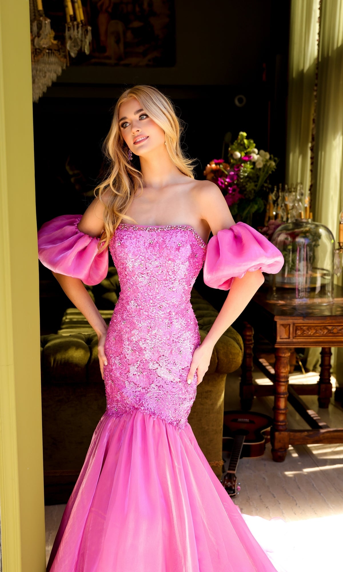 A woman in the Ava Presley 39570 puff-sleeve long prom dress stands indoors near a window, with flowers and a table in the background.