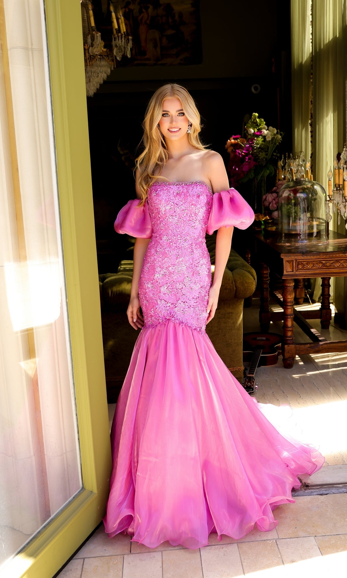A woman stands in a doorway wearing the Ava Presley 39570, a fitted pink mermaid prom dress with off-the-shoulder puff sleeves, embellished bodice, and flared tulle skirt.