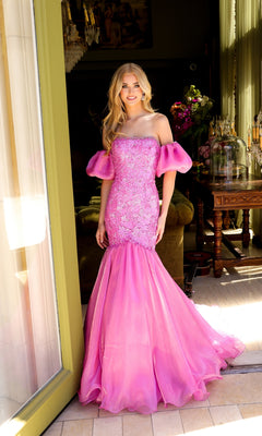 A woman stands in a doorway wearing the Ava Presley 39570, a fitted pink mermaid prom dress with off-the-shoulder puff sleeves, embellished bodice, and flared tulle skirt.
