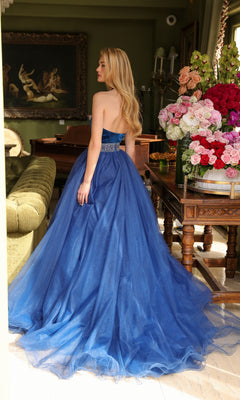 A woman wearing the TRUE a-line evening gown with a beaded waist stands indoors, her back to the camera, near a floral arrangement and antique-style furniture.