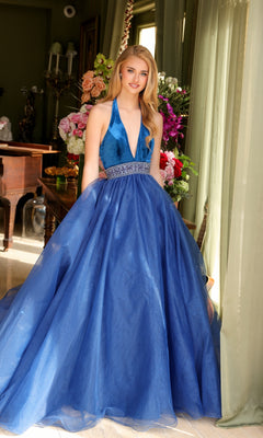 A young woman stands indoors before flowers, wearing TRUE—a blue, floor-length A-line evening gown with a deep V-neck and beaded, fitted waist.