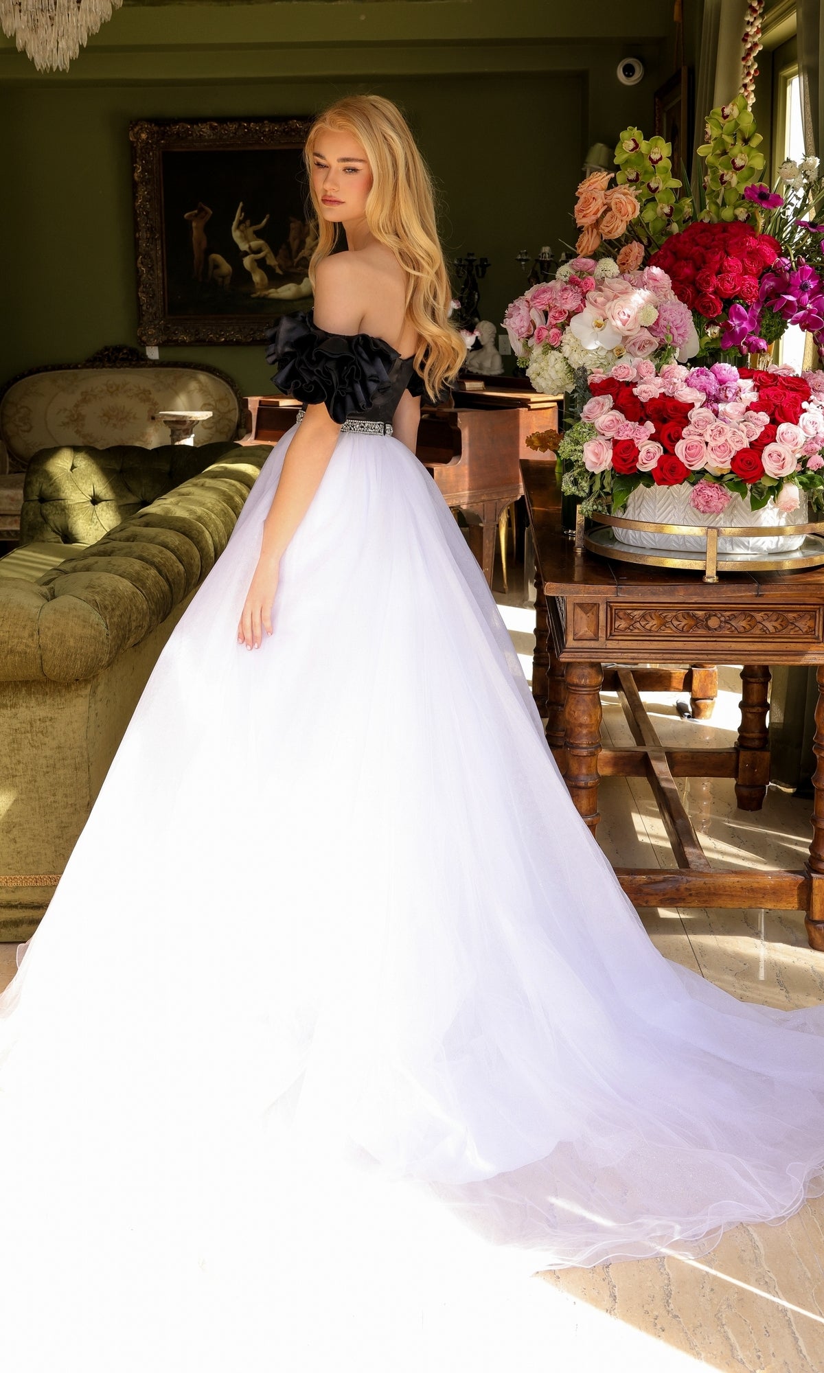 A woman in the Long Prom Dress 39581 by Ava Presley, an off-the-shoulder black and white A-line ball gown, stands indoors near a green sofa and a table with a large bouquet of colorful flowers.