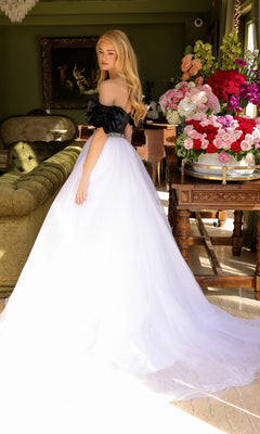 A woman in the Long Prom Dress 39581 by Ava Presley, an off-the-shoulder black and white A-line ball gown, stands indoors near a green sofa and a table with a large bouquet of colorful flowers.