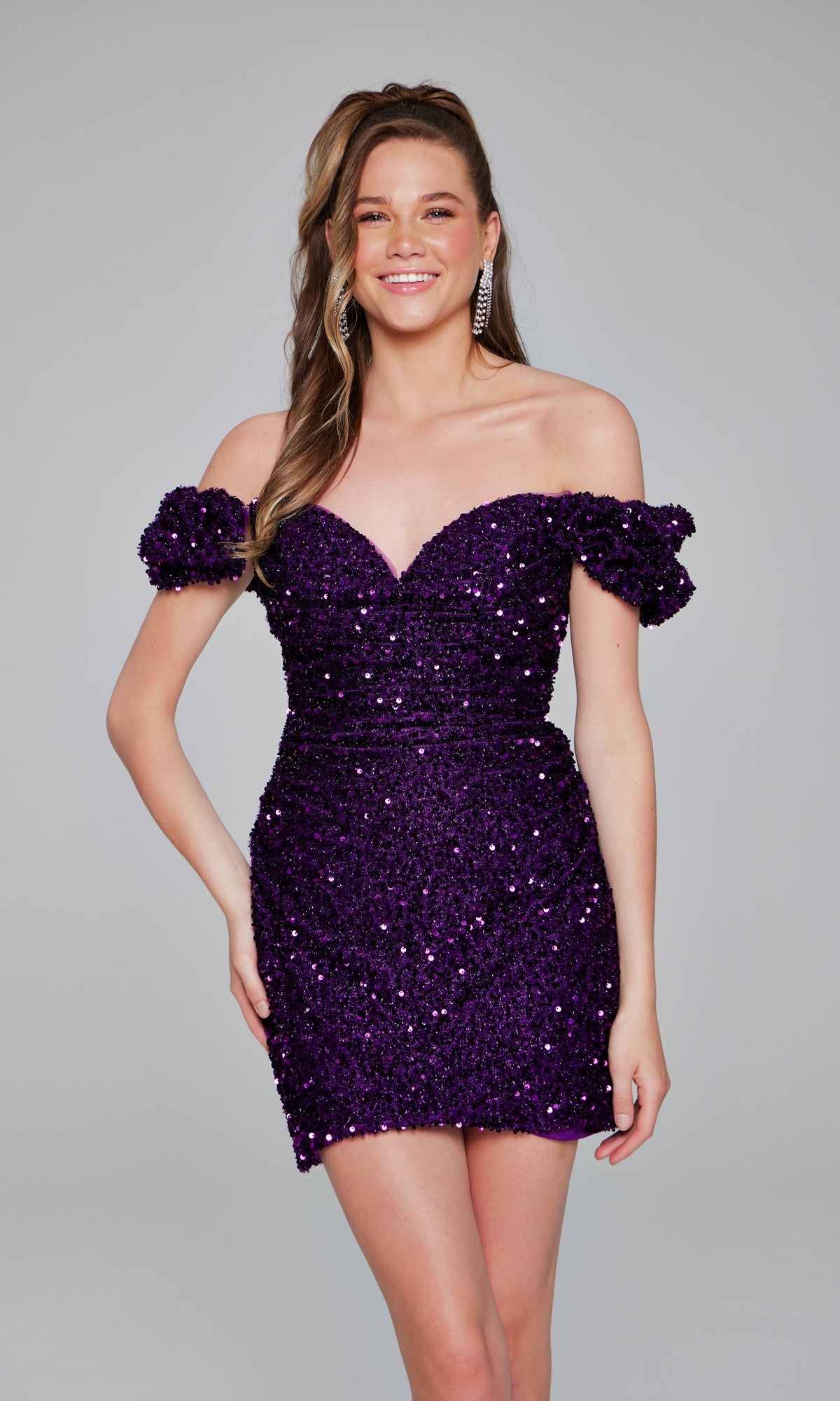 A smiling woman stands against a gray background, wearing the Jovani Off-Shoulder Short Sequin Hoco Dress 39631 in dazzling purple—a sparkling party dress that radiates confidence.