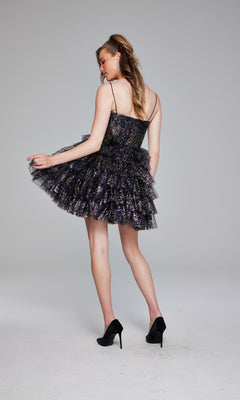 A woman in the Jovani 39654 short black glitter hoco dress and black heels stands with her back turned, lightly lifting the multi-layered, sequined skirt.