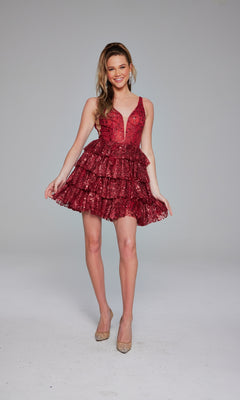 A woman stands smiling against a light background, wearing the Jovani 39700 Ruffled Short Glitter Hoco Dress—a sleeveless deep red, sequined, tiered designer dress—paired with nude heels.