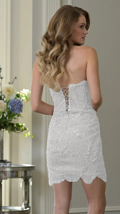 A woman with wavy hair wears the Short Homecoming Dress: Colors Dress 3970—a strapless sequin dress with a lace-up back—standing indoors near a table with flowers, making an elegant choice for homecoming.