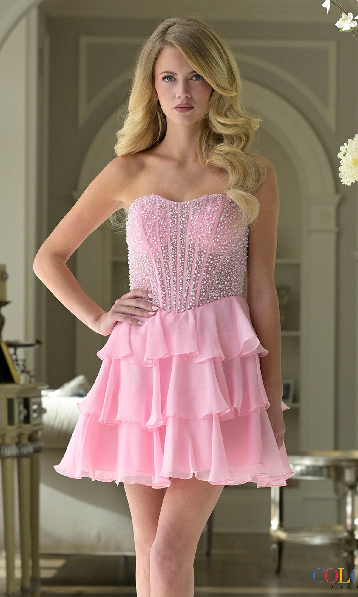 A woman stands indoors wearing the Short Homecoming Dress: Colors Dress 3972 in pink, featuring a strapless design with a beaded bodice and a tiered ruffle chiffon skirt.