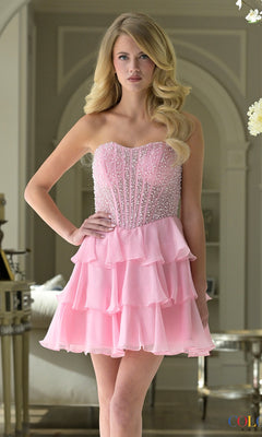 A woman stands indoors wearing the Short Homecoming Dress: Colors Dress 3972 in pink, featuring a strapless design with a beaded bodice and a tiered ruffle chiffon skirt.