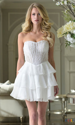 A woman stands indoors near a floral arrangement wearing the Short Homecoming Dress: Colors Dress 3972 in white, featuring a strapless beaded bodice and ruffle chiffon skirt.