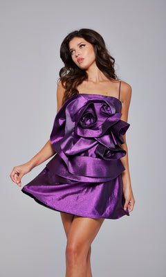 A woman wears the Short Purple Metallic Hoco Dress: Jovani 39749, featuring bold ruffled rose-shaped details, as she poses against a plain gray background—a stunning statement piece for any party.