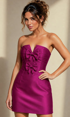A woman wearing the Bow-Front Strapless Short Hoco Dress by Jovani (style 39873) in orchid, featuring a fitted silhouette and two large bows on the bodice, stands against a plain beige background.