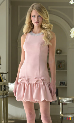 Woman wearing Colors Dress 3991, a short pink sleeveless sheath homecoming dress with bow accents and ruffled hem, standing indoors by decorative shelves—a charming party look.