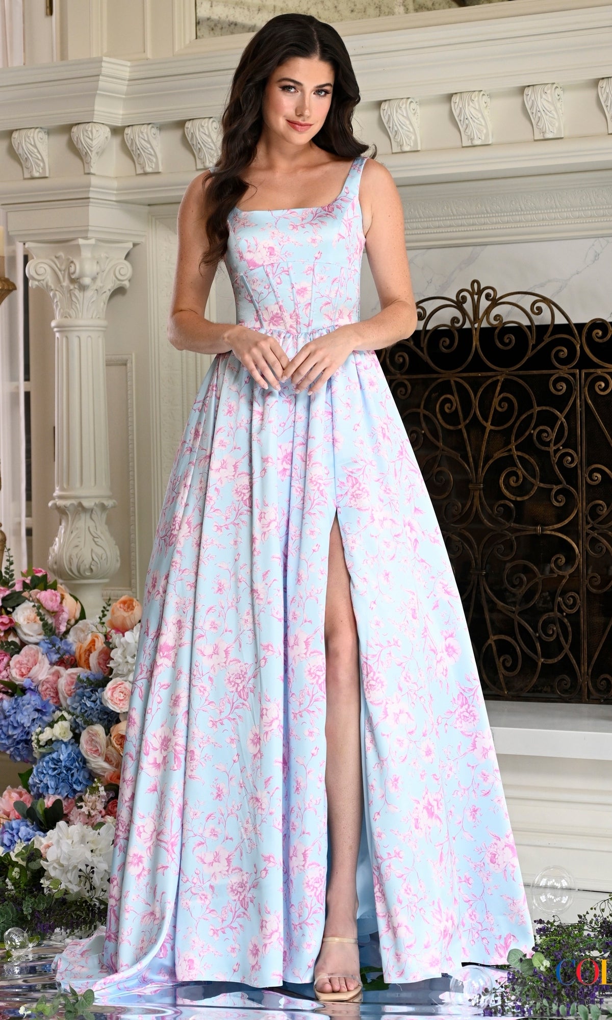 A woman wearing a Long Prom Dress: Colors Dress 3996 in blue and pink, featuring a floor-length floral pattern and a front thigh-high slit.