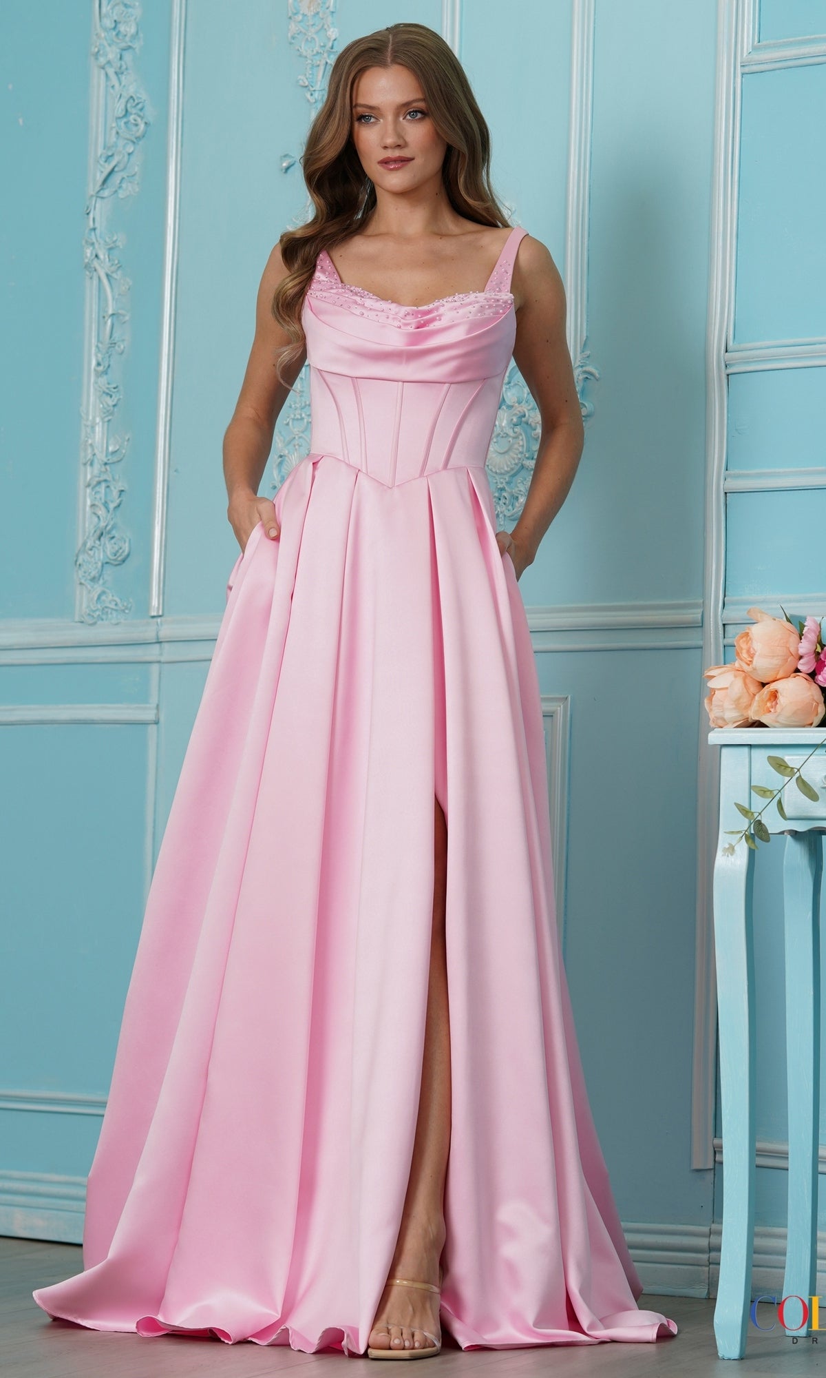 A woman wearing a pink satin Colors Dress 4001 prom dress with a cowl neckline, corset-style bodice, and a high leg slit.