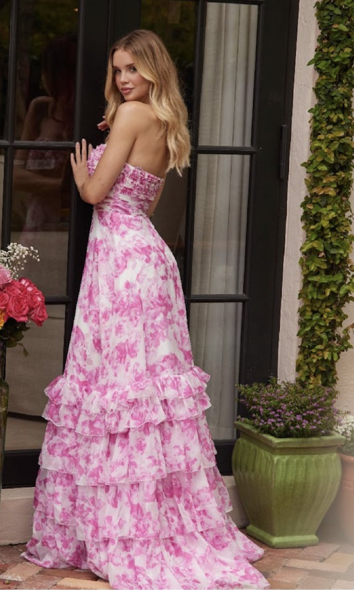 A woman in the Ava Presley 40026 Keyhole Floral-Print Prom Dress stands by glass doors amid flowers and greenery, her strapless tiered a-line skirt with pink and white floral print flowing gracefully to the floor.
