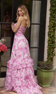 A woman in the Ava Presley 40026 Keyhole Floral-Print Prom Dress stands by glass doors amid flowers and greenery, her strapless tiered a-line skirt with pink and white floral print flowing gracefully to the floor.