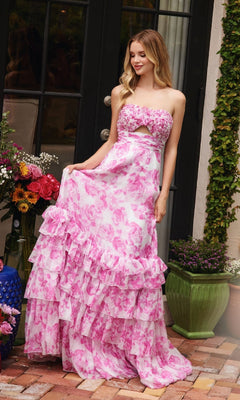 A woman stands on a brick patio near potted plants, wearing the Ava Presley 40026 Keyhole Floral-Print Prom Dress—a strapless, pink and white gown with a tiered A-line skirt—holding the skirt by a glass door.