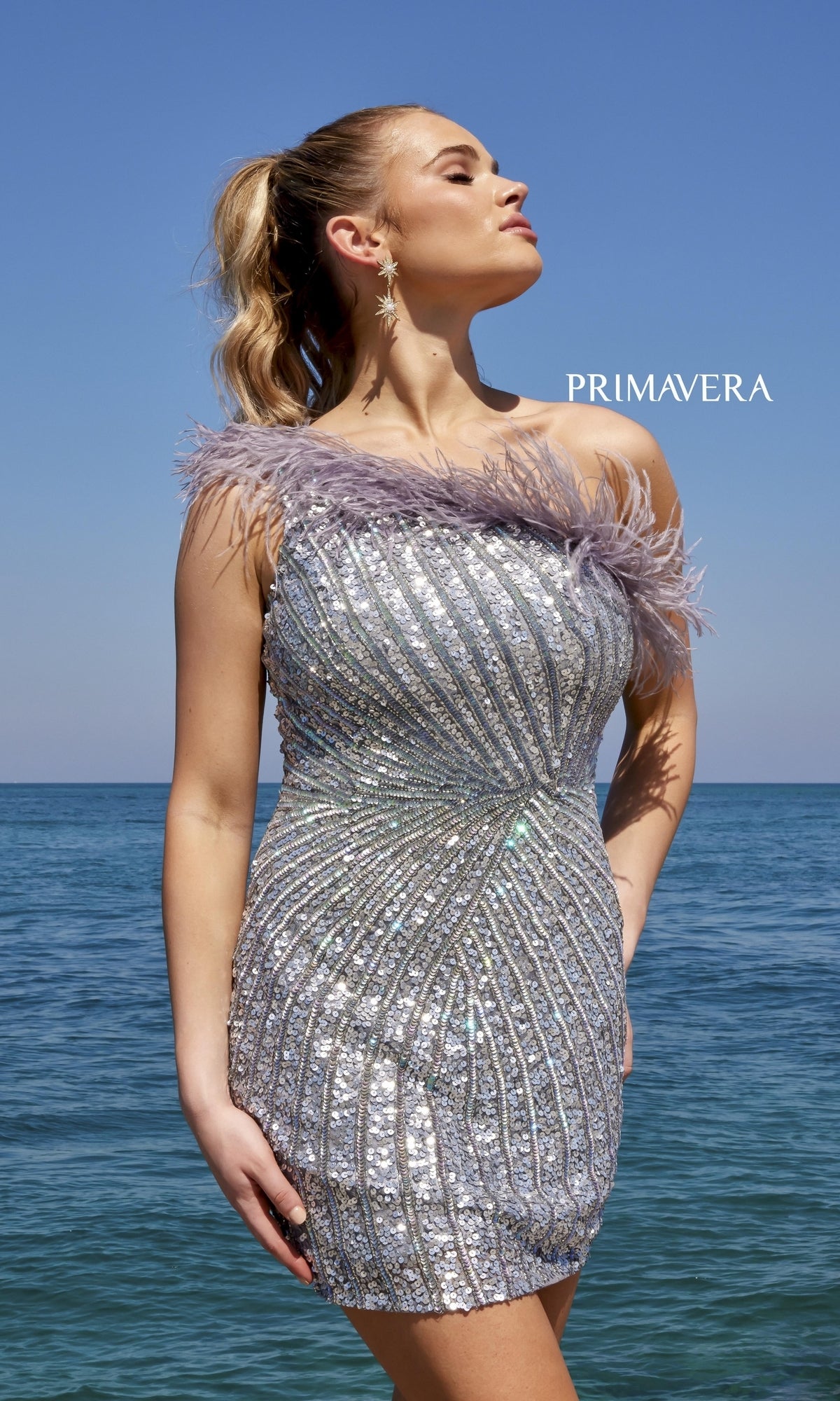 A woman wearing the One-Shoulder Sequin Party Dress with Feathers 4002 stands by the sea under a clear blue sky. The word 