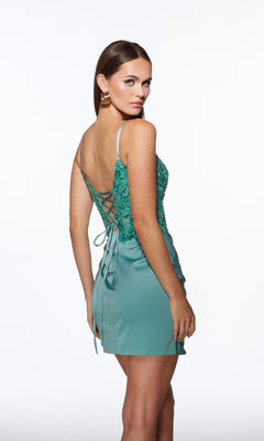 Woman wearing the Alyce 40036 Sheer-Corset Short Homecoming Dress with a teal, sleeveless bodycon fit, floral details, and lace-up back, standing and looking over her shoulder against a plain white background.