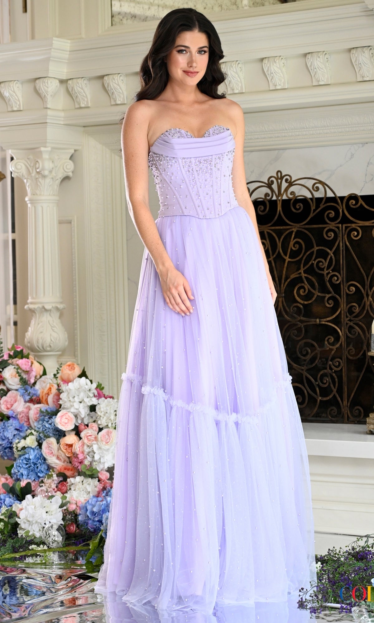 A woman wears a strapless, lilac-colored Long Prom Dress: Colors Dress 4003, featuring a corset-style bodice with beaded embellishments and a layered tulle skirt.