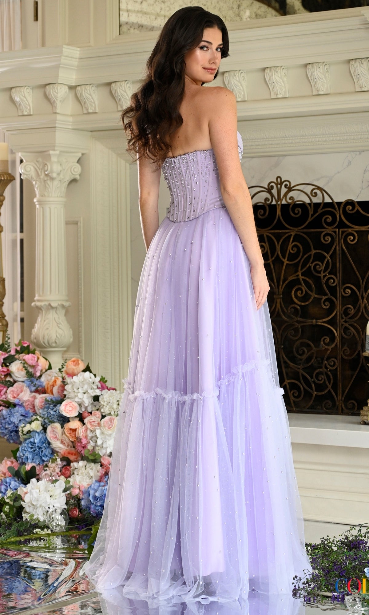 A woman wears a lavender strapless tulle Colors Dress 4003 prom dress with a beaded corset bodice and a tiered floor-length skirt.