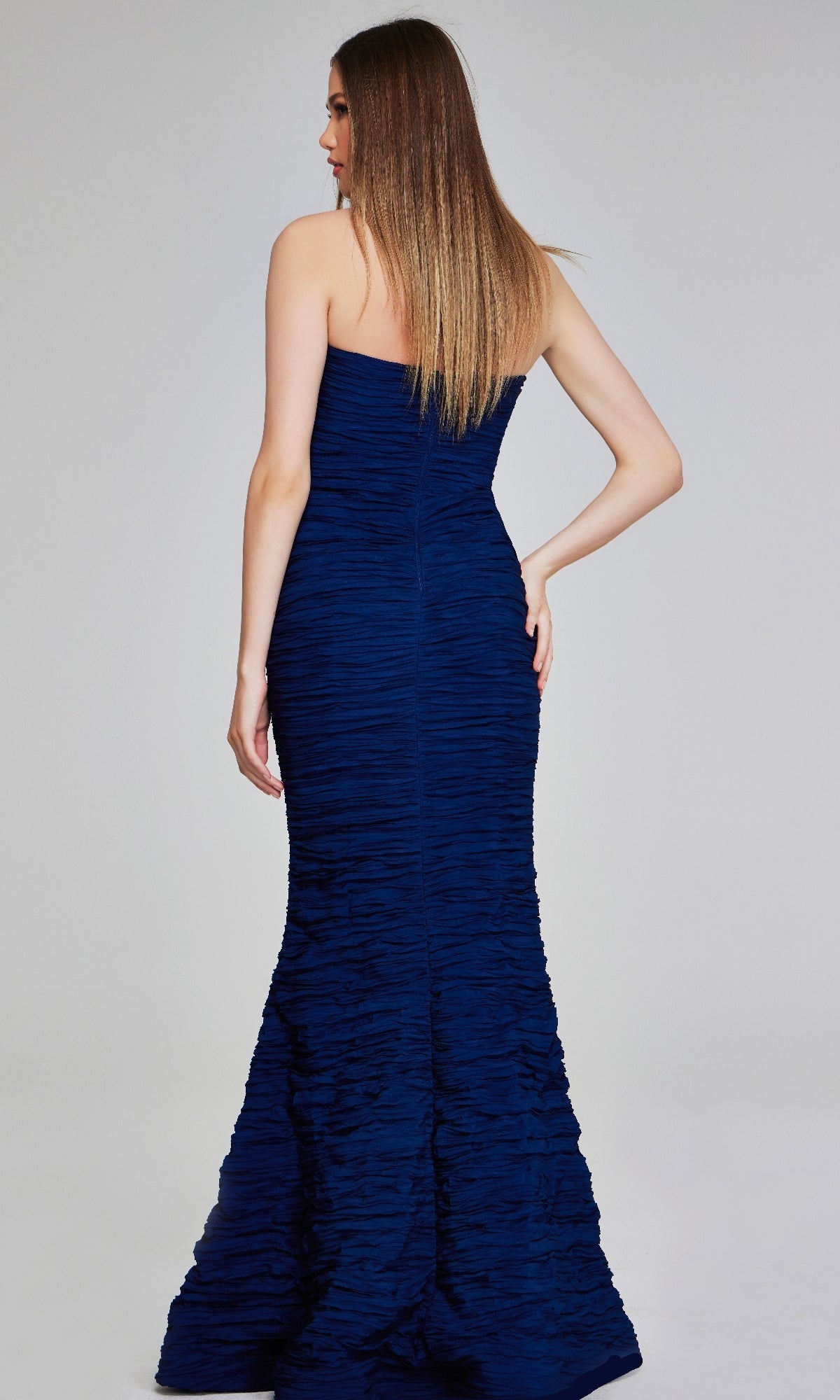 A woman stands facing away in the Jovani 40045, a strapless deep blue mermaid evening gown with elegant ruching, set against a plain light background.