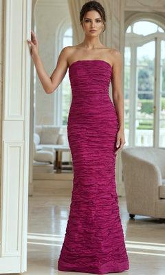 A woman stands indoors near a white wall, with elegant furniture and large windows in the background, wearing the Long Formal Dress: Jovani 40045—a strapless, ruched evening gown in textured magenta.