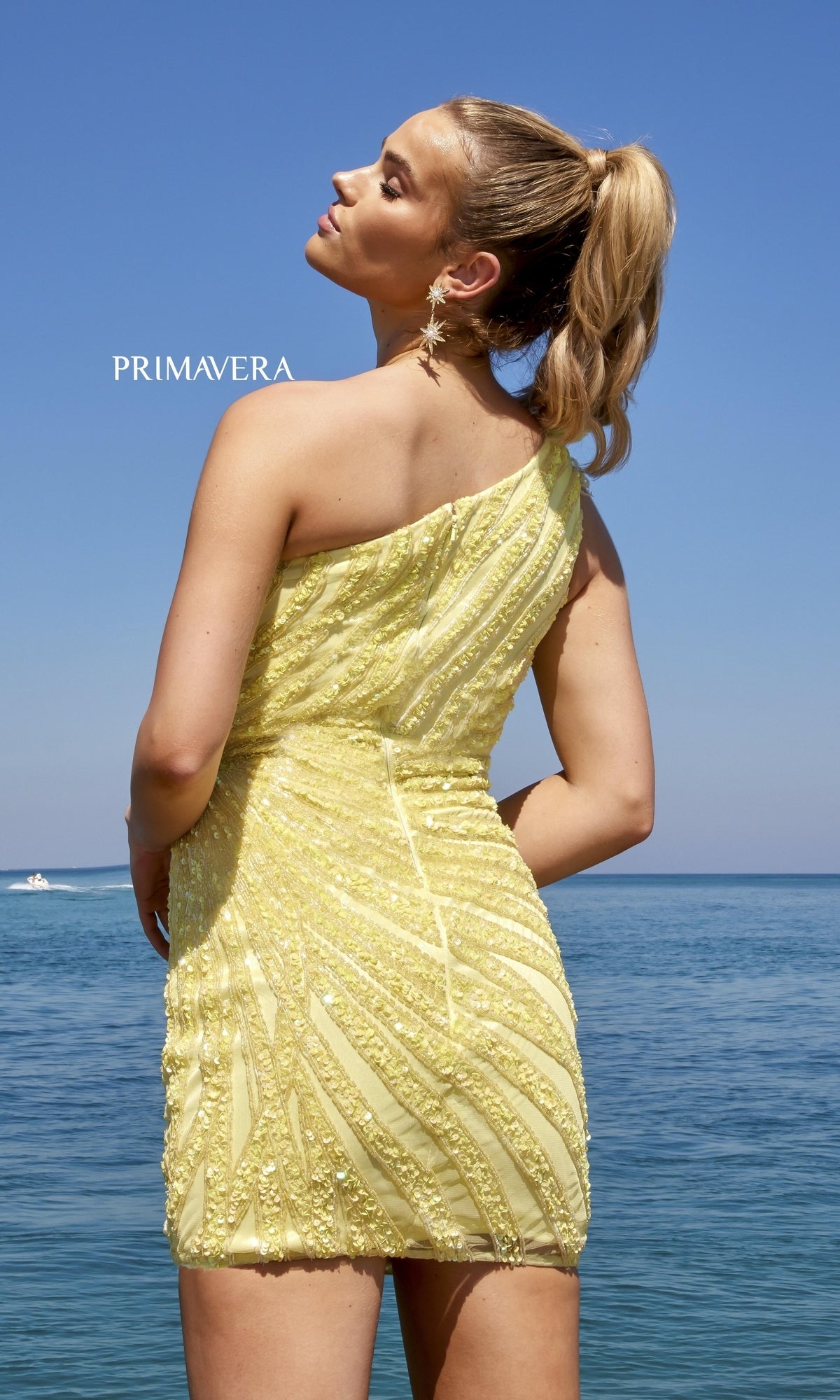 A woman in the One-Shoulder Feathered Beaded Cocktail Dress 4004, yellow and beaded, stands by the ocean with her hair in a ponytail under a clear blue sky.