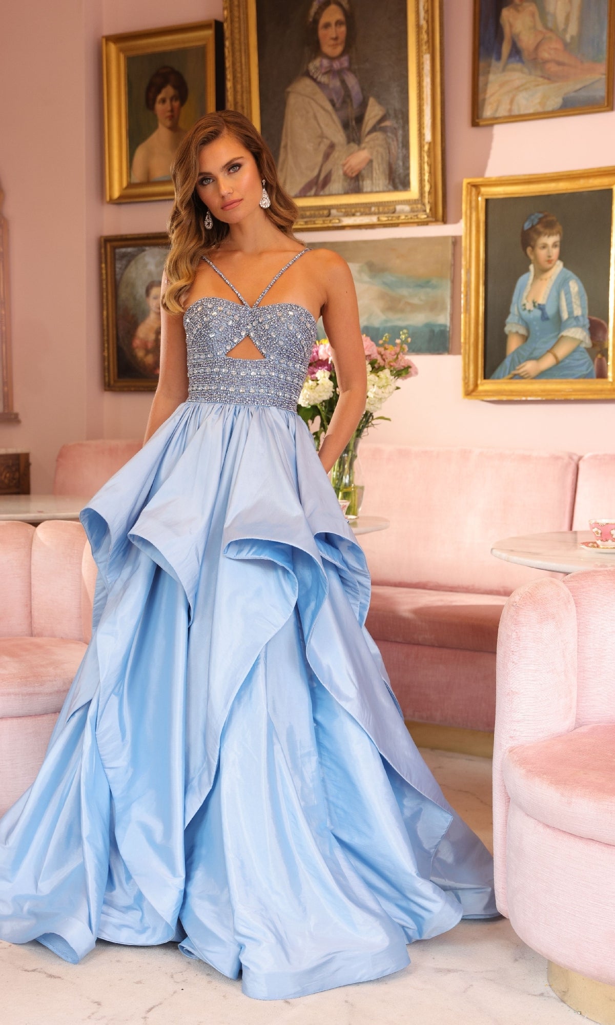 A woman wears the sparkly light blue Horsehair-Hem Prom Ball Gown: Ava Presley 40054 in a pink room with framed portraits and a pink sofa in the background.