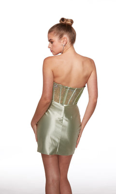A woman stands with her back to the camera wearing the Alyce 40054 short homecoming dress in light green, featuring a strapless, beaded rhinestone corset bodice and a fitted satin skirt.