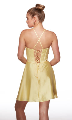 Woman faces away, wearing the Alyce 40063 Short Satin Homecoming Dress in yellow, featuring crisscross straps and lace-up back details, standing against a white background.
