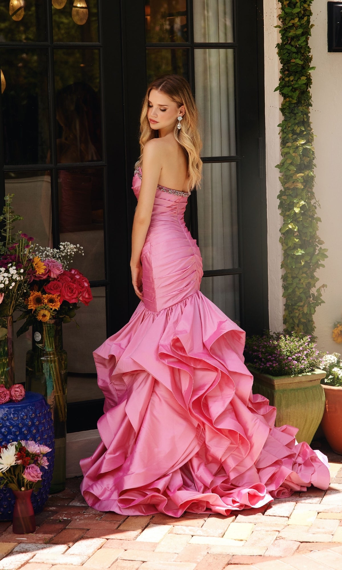 A woman in the Ava Presley 40068 Strapless Ruffled Mermaid Prom Dress, featuring a pleated bodice and ruffled mermaid skirt, stands on a brick patio near a glass door, surrounded by potted plants and colorful flowers.