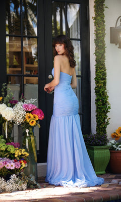 A woman wearing the Ava Presley 40073 Strapless Ruched Mermaid Prom Dress stands outdoors by glass doors, surrounded by vibrant flower arrangements.