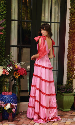 A woman wearing the Shoulder Bows A-Line Formal Dress: Ava Presley 40091 stands on a brick patio before a glass door, surrounded by colorful flower arrangements in assorted vases.